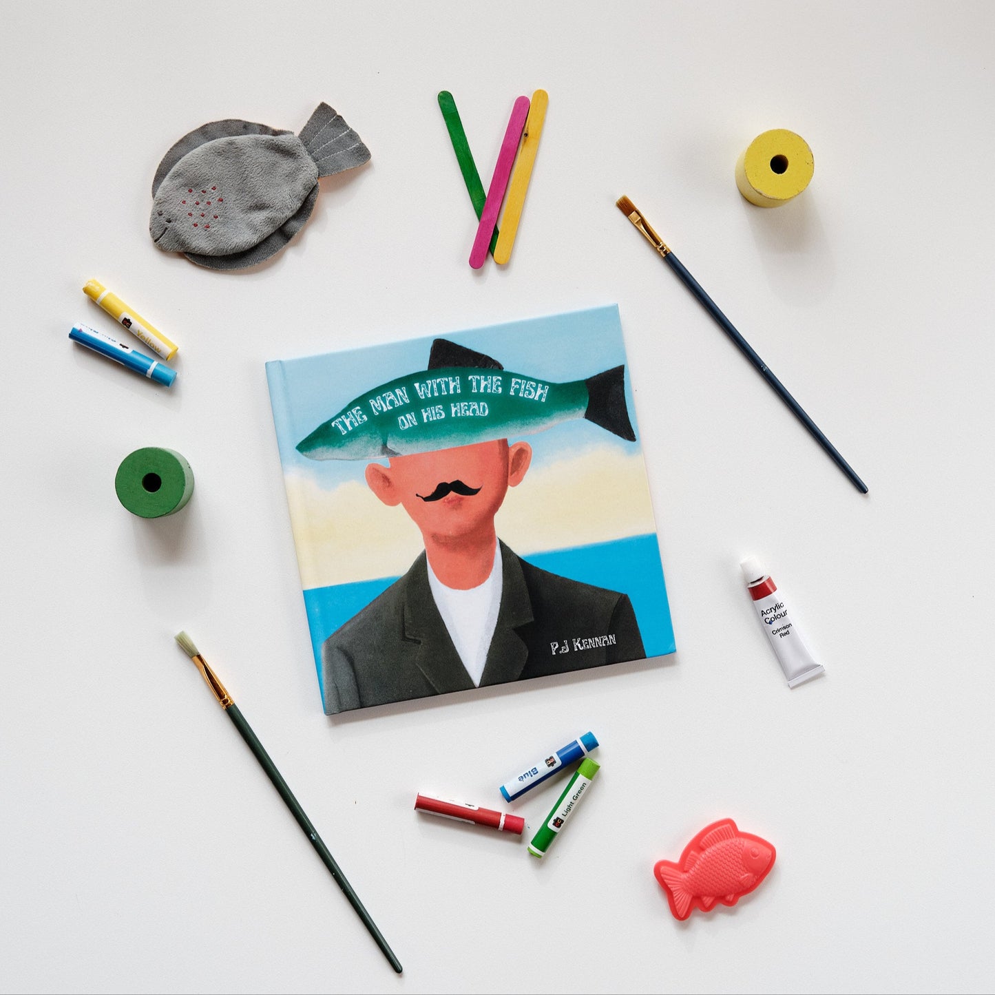 Image of book on white background surrounded with objects such as plush fish, paint brushes, paint and crayons